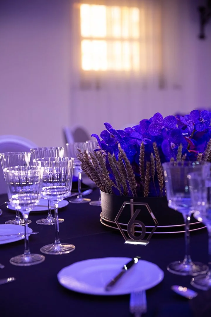 Eleganter festlich gedeckter Tisch mit violetter Blumenarrangement, leeren Weingläsern, weißen Tellern und silbernem Besteck bei Abendveranstaltung.