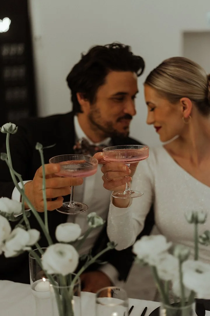 Ein Mann und eine Frau feiern einen besonderen Anlass, sie stoßen mit rosa Cocktails an, im Vordergrund sind weiße Blumen und Kerzen zu sehen.