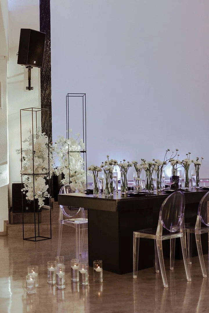Eine festlich gedeckte Tafel mit weißen Blumen in Gläsern, Kerzen in Gläsern auf dem Boden, transparente Stühle, und dekorative metallene Rahmen, wahrscheinlich für eine Veranstaltung oder eine Feier.