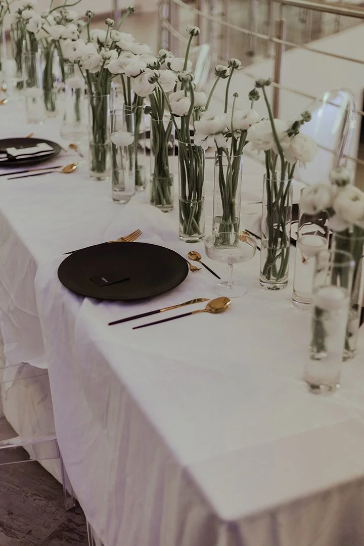 Eleganter Tisch mit weißen Blumen in hohen Gläsern, schwarzem Teller, Gold- und schwarzen Besteckteilen, Glas Wasser, und gedeckten Sitzplätzen bei einer Veranstaltung.