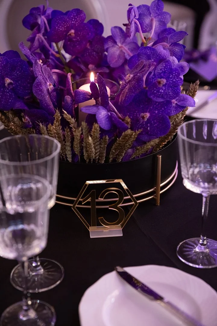 Tischdeko mit violetten Blumen, Kerze, Zahl 13 in Gold auf Tisch, Besteck und Gläser.