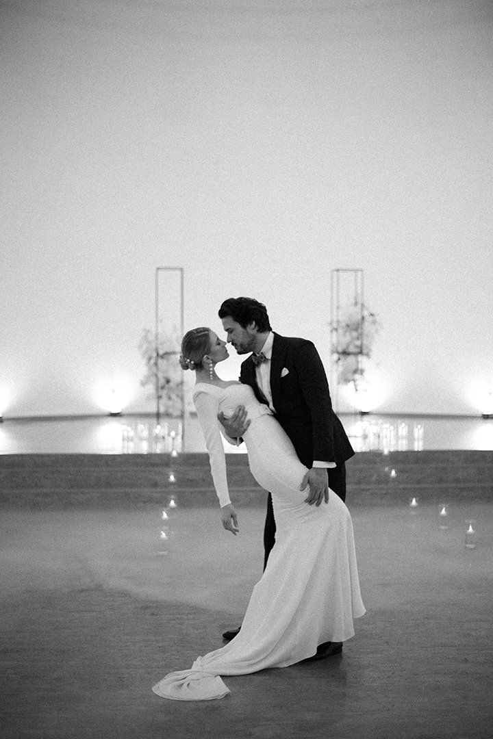 Ein Paar in formeller Kleidung tanzt bei einer Hochzeit oder Gala, die Frau trägt ein langes, weißes Kleid und der Mann einen schwarzen Anzug, in einem minimalistischen, eleganten Raum.