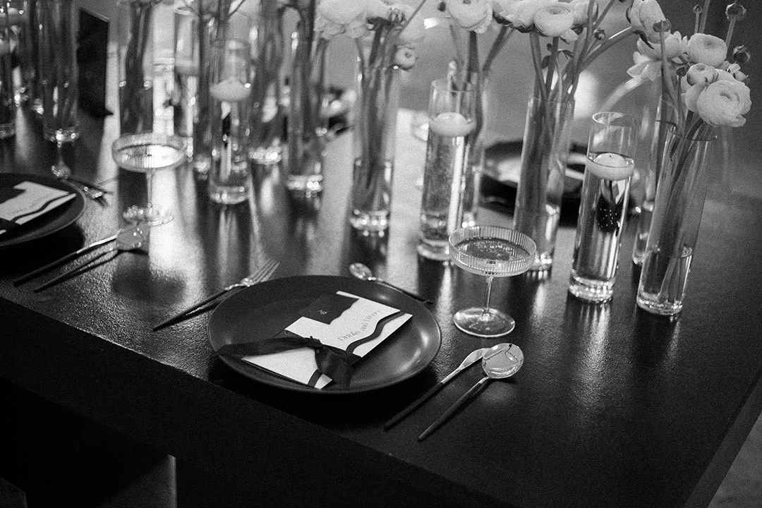 Eleganter gedeckter Tisch mit schwarzen Tellern, Besteck, Gläsern und weißen Blumen in Vasen, schwarz-weißes Foto.