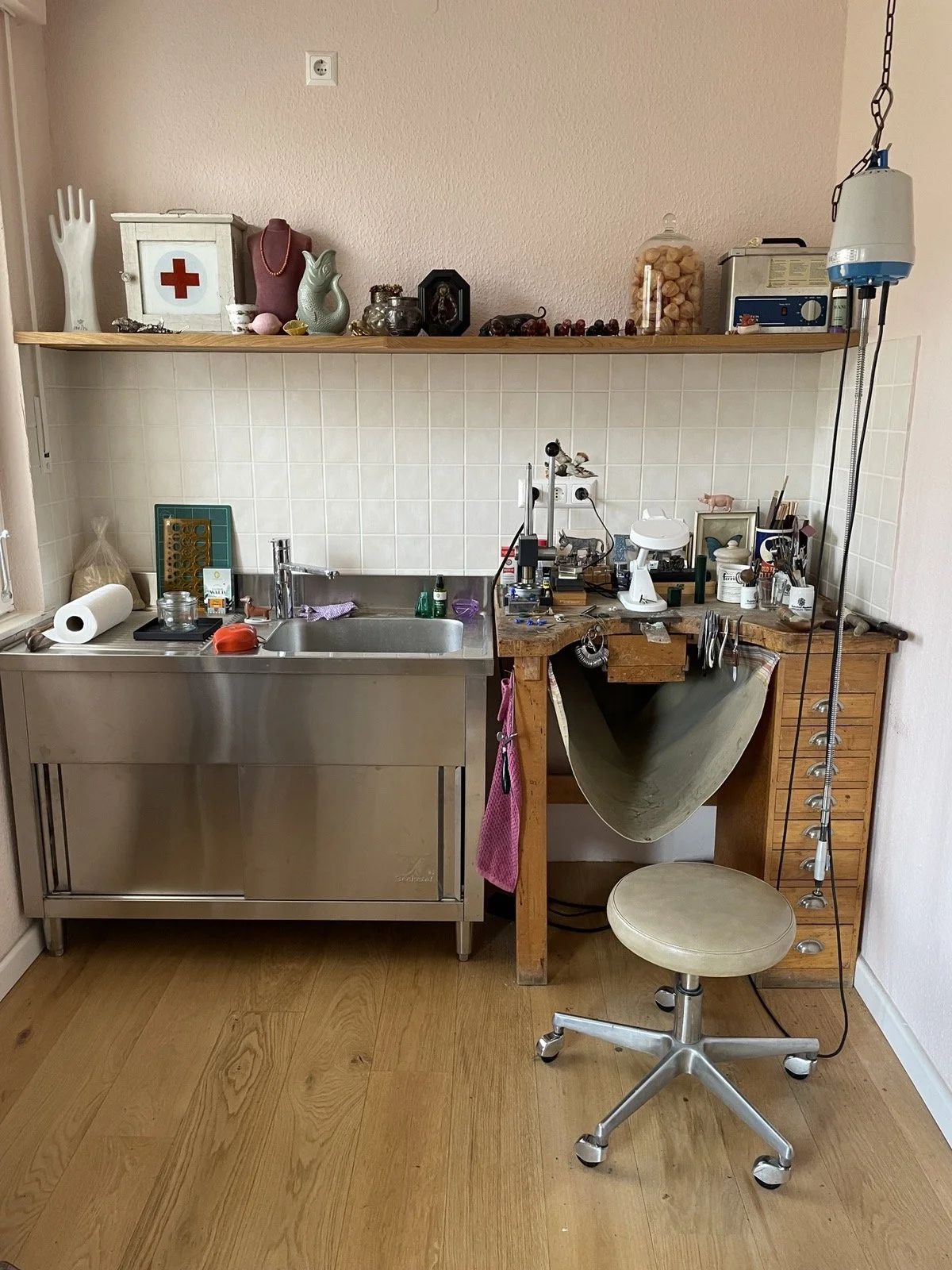Arbeitsplatz für Handarbeit mit Werkzeugen, Maschinen, Stuhl und verschiedenen Gegenständen, darüber eine offene Holzablage mit Dekogegenständen, medizinischen und Küchenartikeln, in einem Raum mit Holzboden.