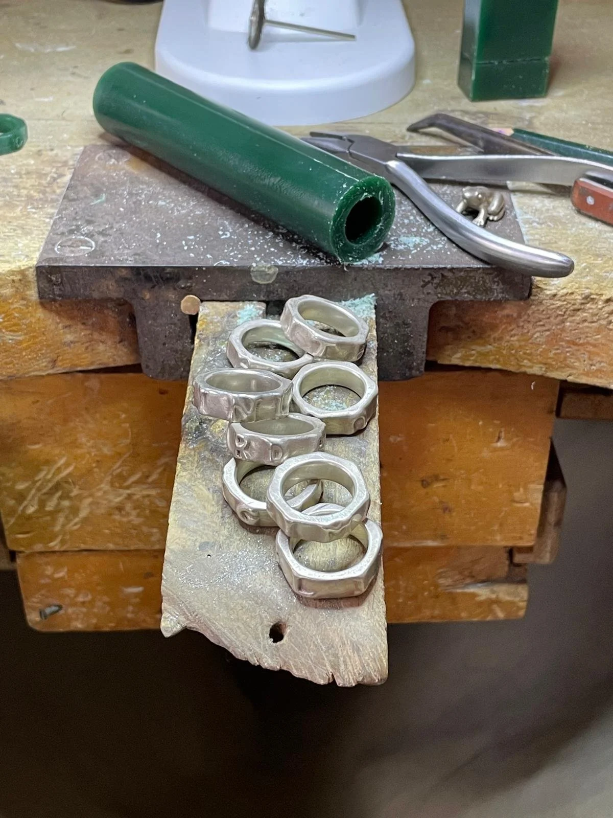Silberne Ringe auf einem werkbankähnlichen Tisch, ein grüner Rohling, verschiedene Werkzeuge und ein Stück Holz.