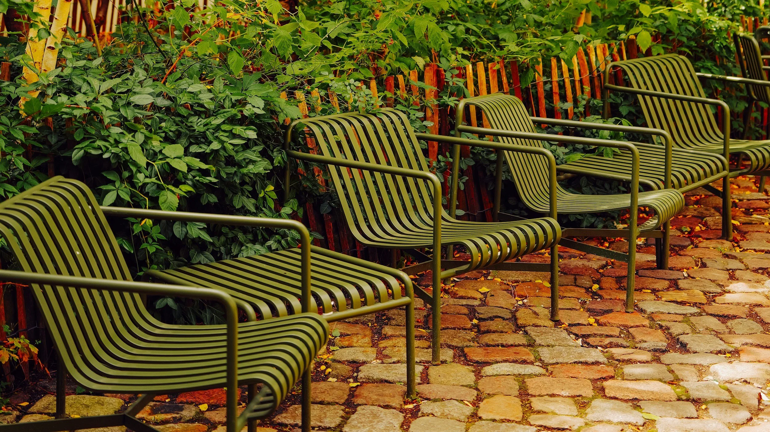 Several green metal benches with striped seats and backs arranged along a cobblestone path, with green bushes and a wooden fence in the background.