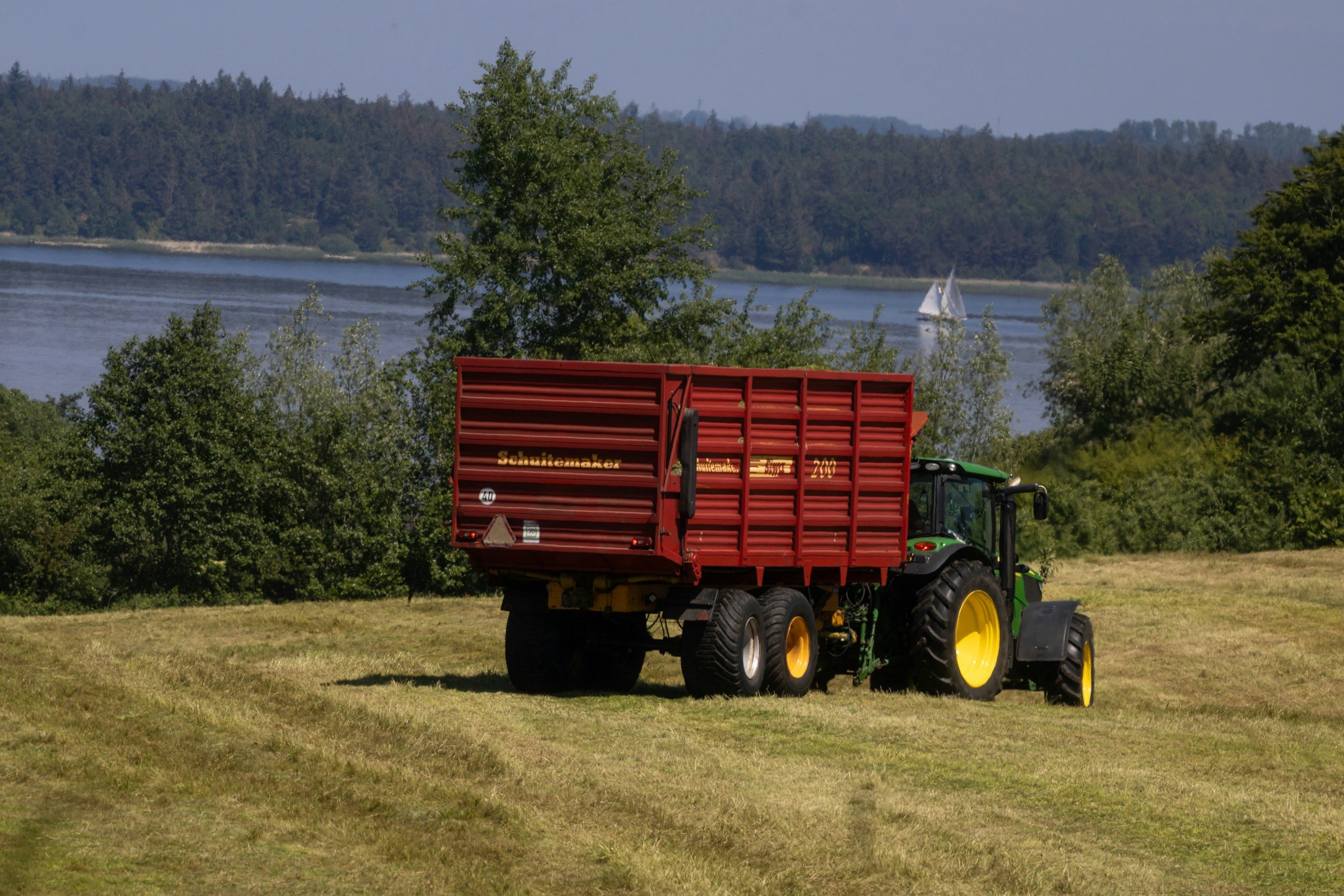 A green tractor pulling a red trailer with yellow wheels parked on a grassy field near a lake, with trees and sailboats visible in the background.