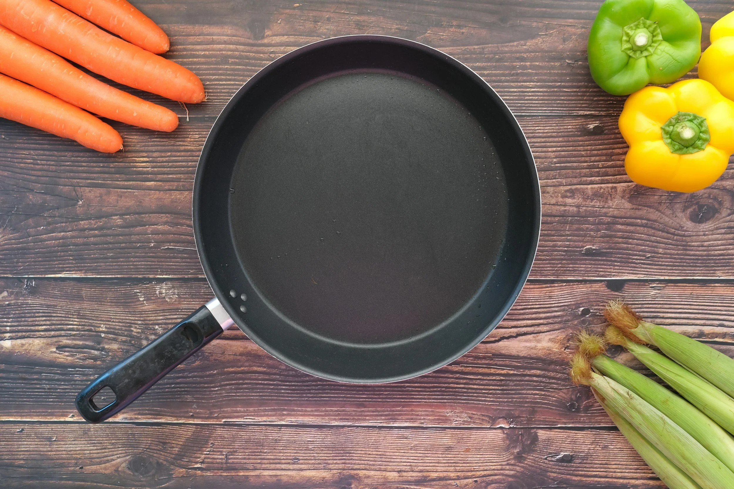 A black frying pan on a wooden surface with fresh carrots, green bell pepper, yellow bell pepper, and celery.