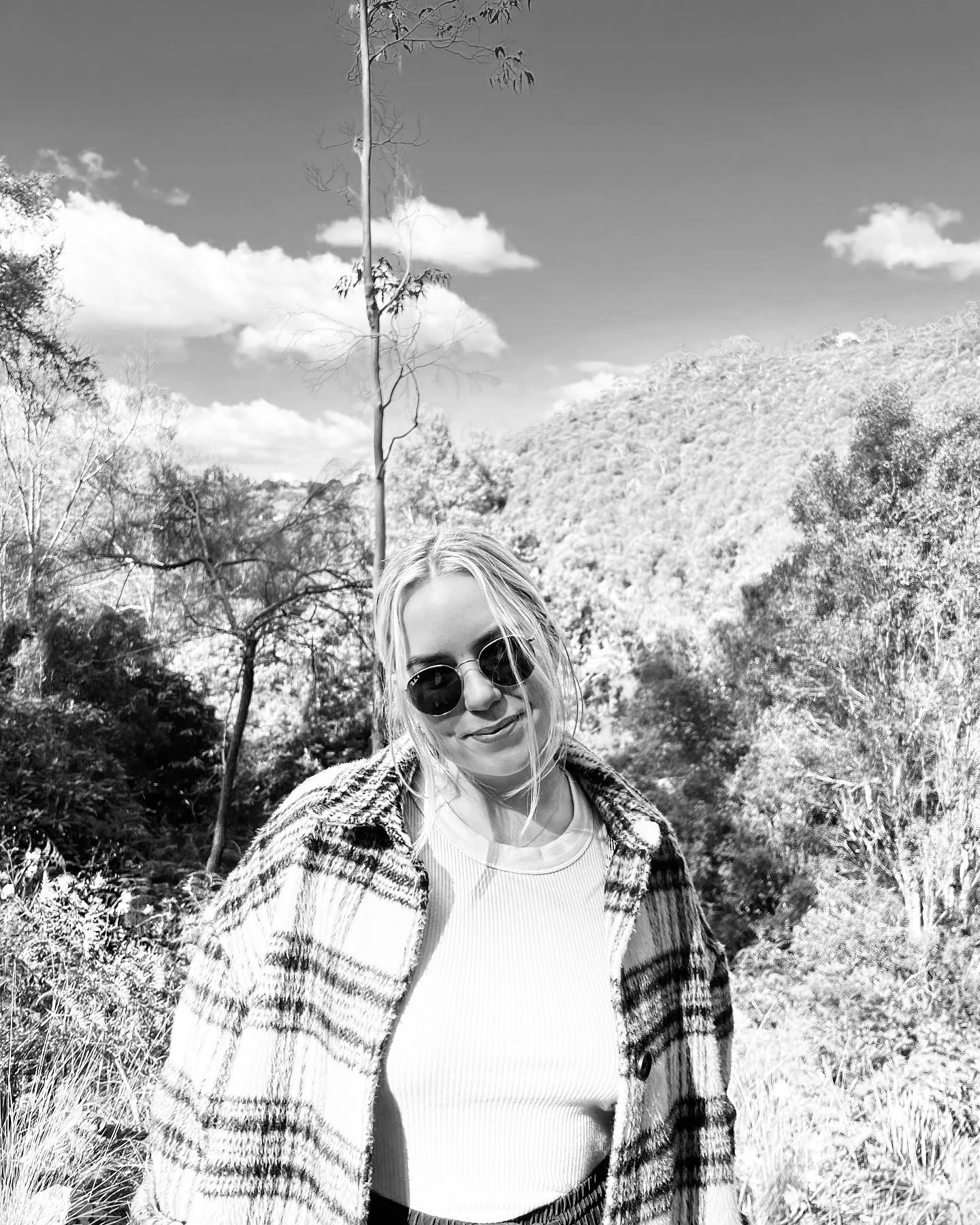 A woman with blonde hair wearing sunglasses and a plaid coat, standing outdoors in a natural landscape with trees, hills, and a partly cloudy sky.
