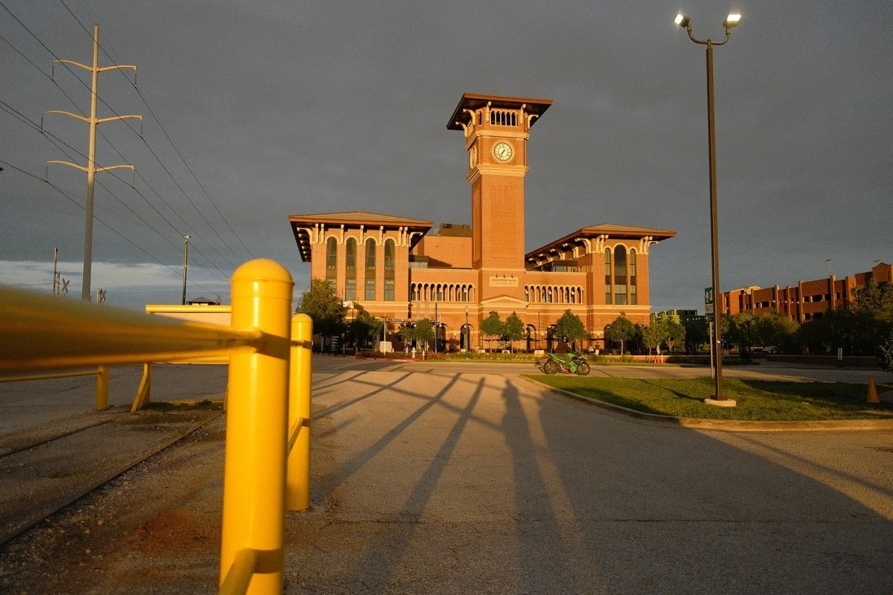 A large historic brick train station with a clock tower, surrounded by trees and a parking lot, during sunset with dark clouds overhead.