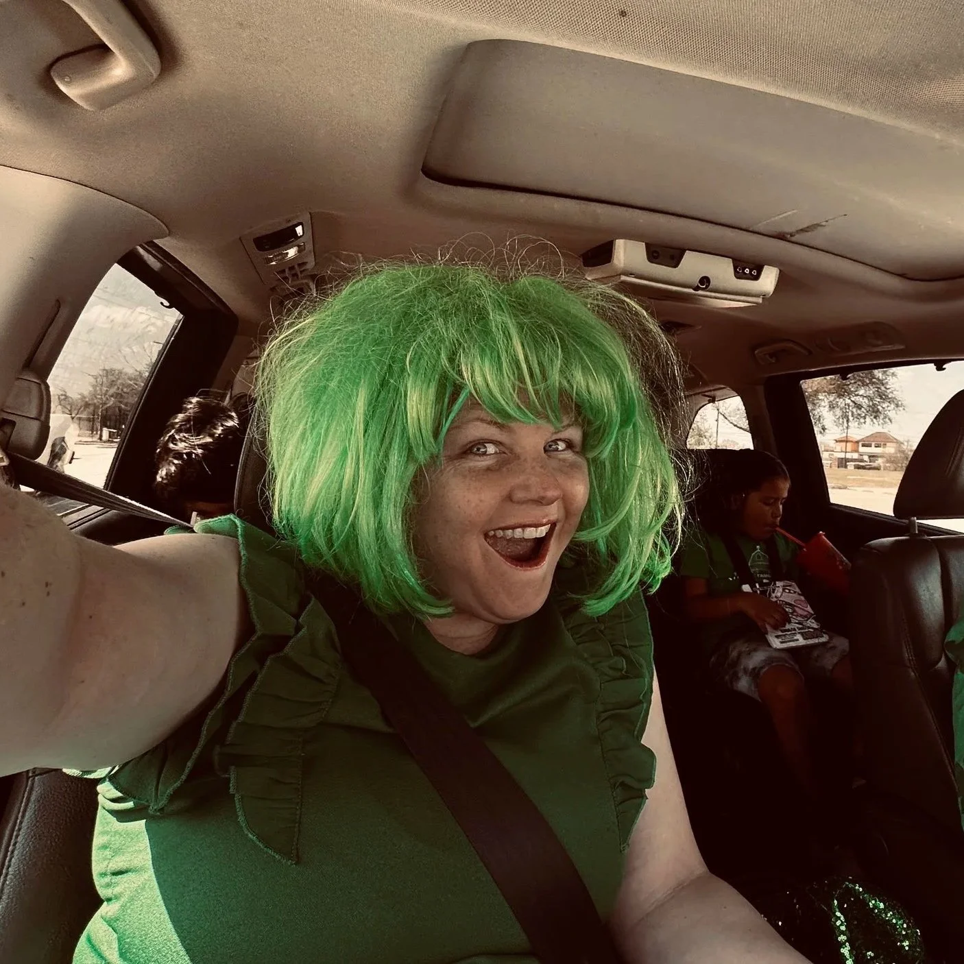 A woman wearing a bright green wig is taking a selfie inside a vehicle, with a cheerful expression on her face. In the background, a girl is sitting in a car seat, looking down at a book or tablet.