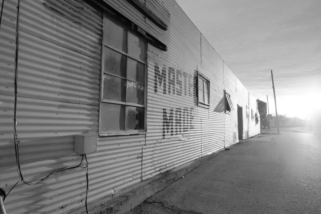A black and white photo of a rundown building with corrugated metal siding and multiple windows. The words 'MOTEL' and 'VACANCY' are spray-painted on the wall. The setting appears to be in a rural or deserted area with an empty street and utility poles.