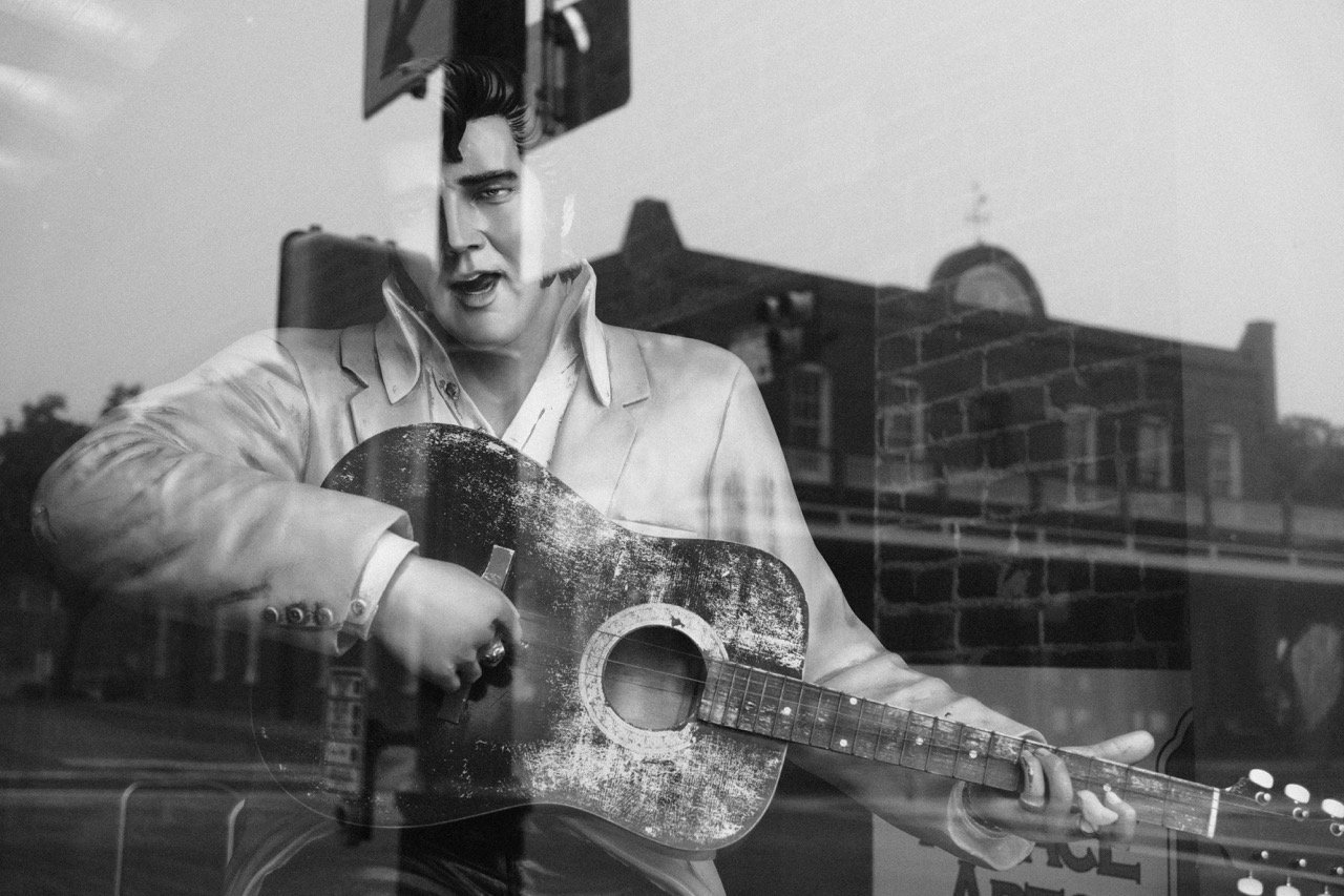 A black and white reflection of a man playing an acoustic guitar through a window. The man appears to be singing, with a building and sky reflected in the glass.