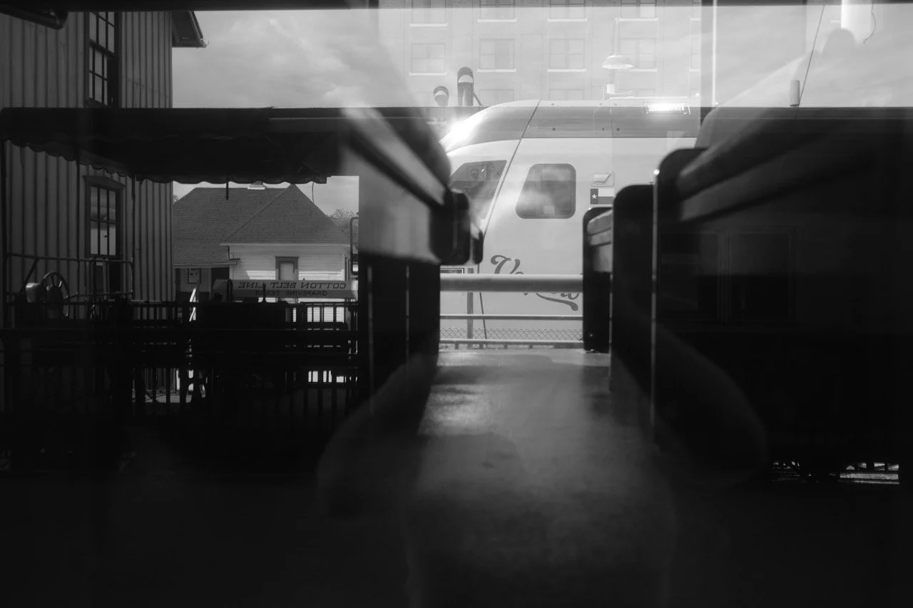 Photo taken from inside a building through a window showing a train outside on the tracks, with some passenger seats visible in the foreground. The scene is in black and white.
