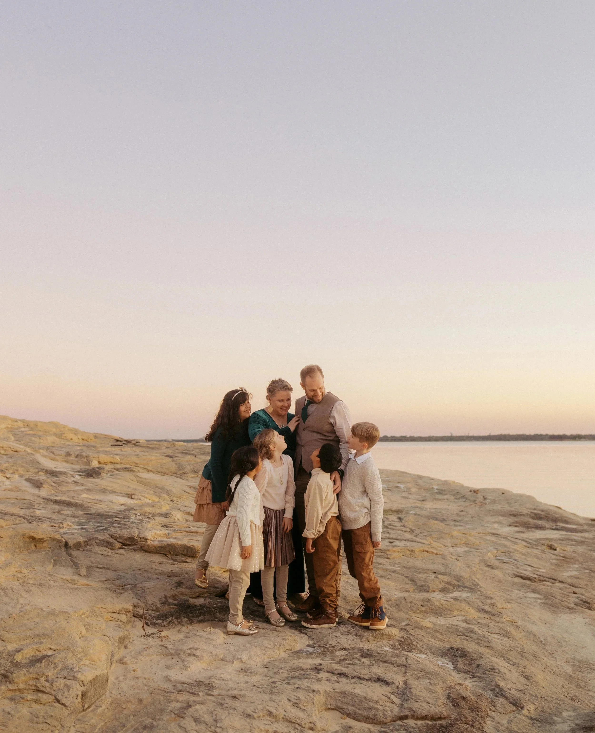Family of seven, including adults and children, gathered together on rocky terrain by water at sunset, smiling and enjoying each other's company.