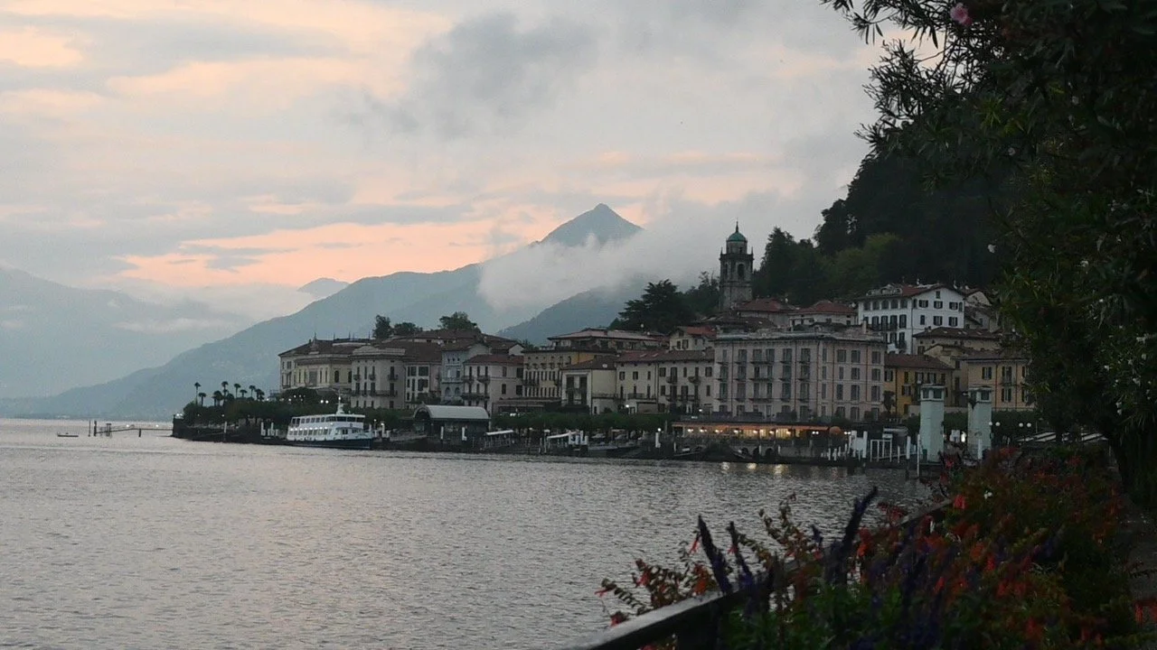 View of a lakeside town with colorful buildings, boat docked at the pier, and mountains in the background during sunset.