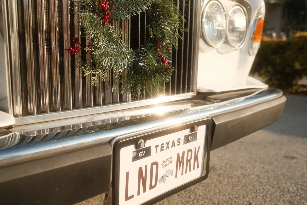 Front of a vintage vehicle decorated with Christmas greenery and red berries, showing a Texas license plate reading 'LND MRK'.