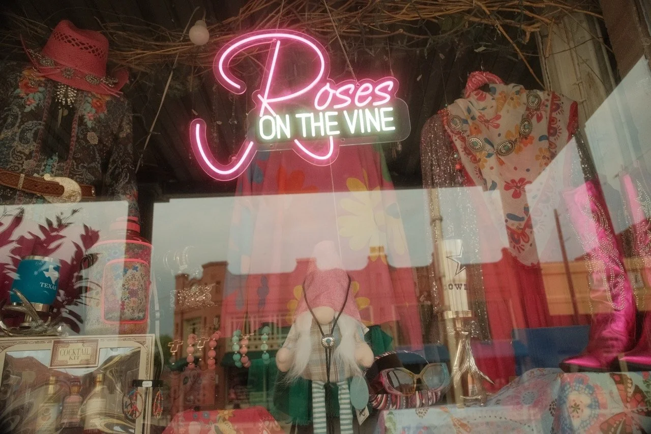 Display window with vintage clothing and accessories, including a pink hat, dresses, jewelry, sunglasses, and decorative items, with a neon sign reading 'Roses on the Vine'.