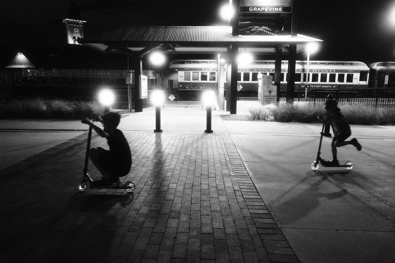 Two children riding scooters at a train station at night with a train in the background.