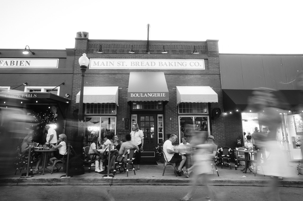 Black and white photo of a street scene in front of a bakery called "BOULANGERIE" with outdoor seating, people dining and walking, and a brick building with signs, awnings, and a streetlamp.