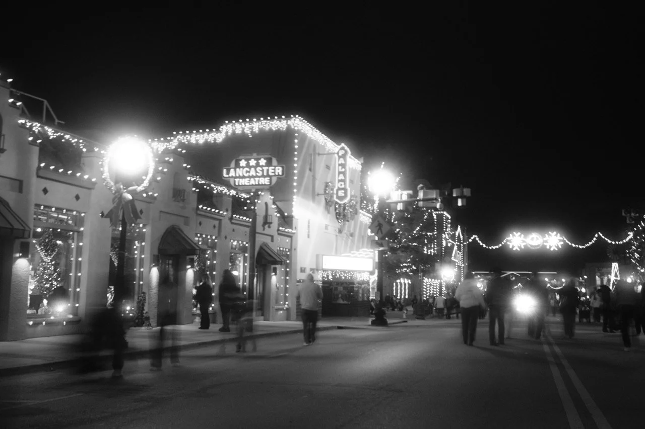 The street scene at night in downtown Lancaster with holiday lights, trees, and buildings decorated for Christmas, including the Lancaster Theatre and Palace sign.