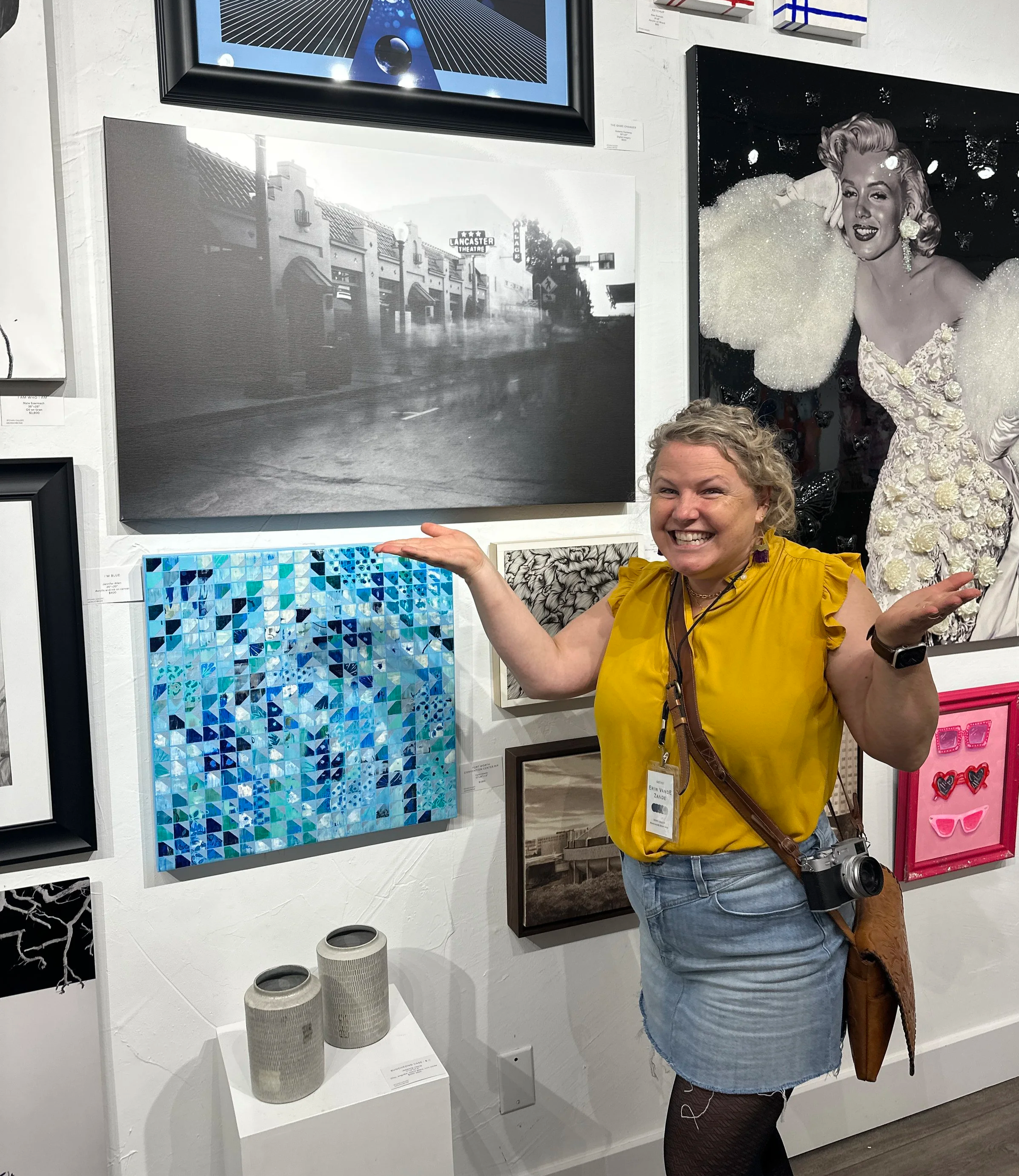 A woman in a yellow top and denim skirt smiling and gesturing at a black and white photo of a street scene on a gallery wall, surrounded by other colorful and black-and-white artwork.