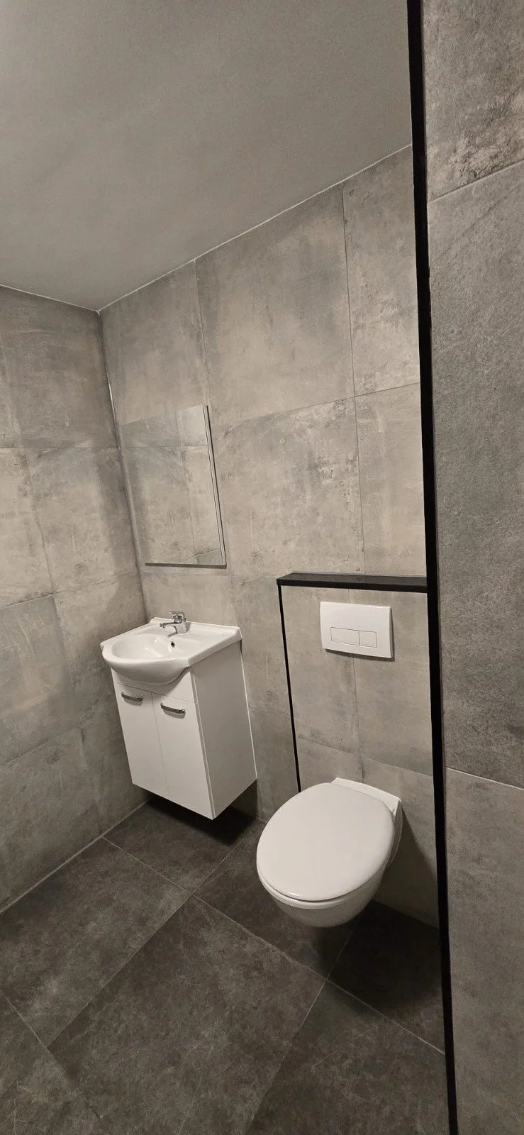 Moderne kleine badkamer met grijze tegelwanden en -vloeren, een wit hangtoilet en een klein wasbakje met onderkast.