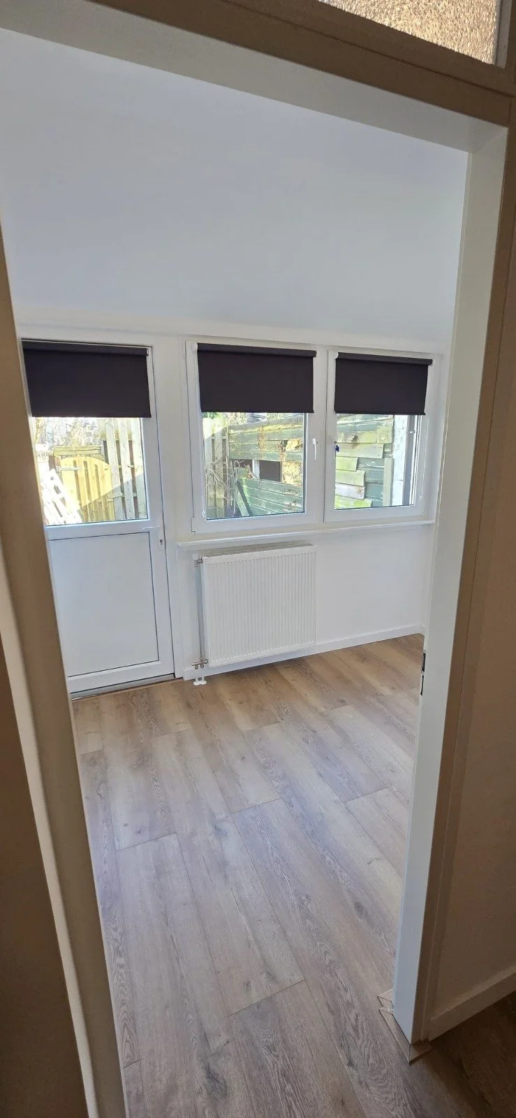 Een kamer met houten vloer, witte muren, drie grote ramen met zwarte rolgordijnen, en een deur met een raam die uitkijken op een tuin met een houten schutting.