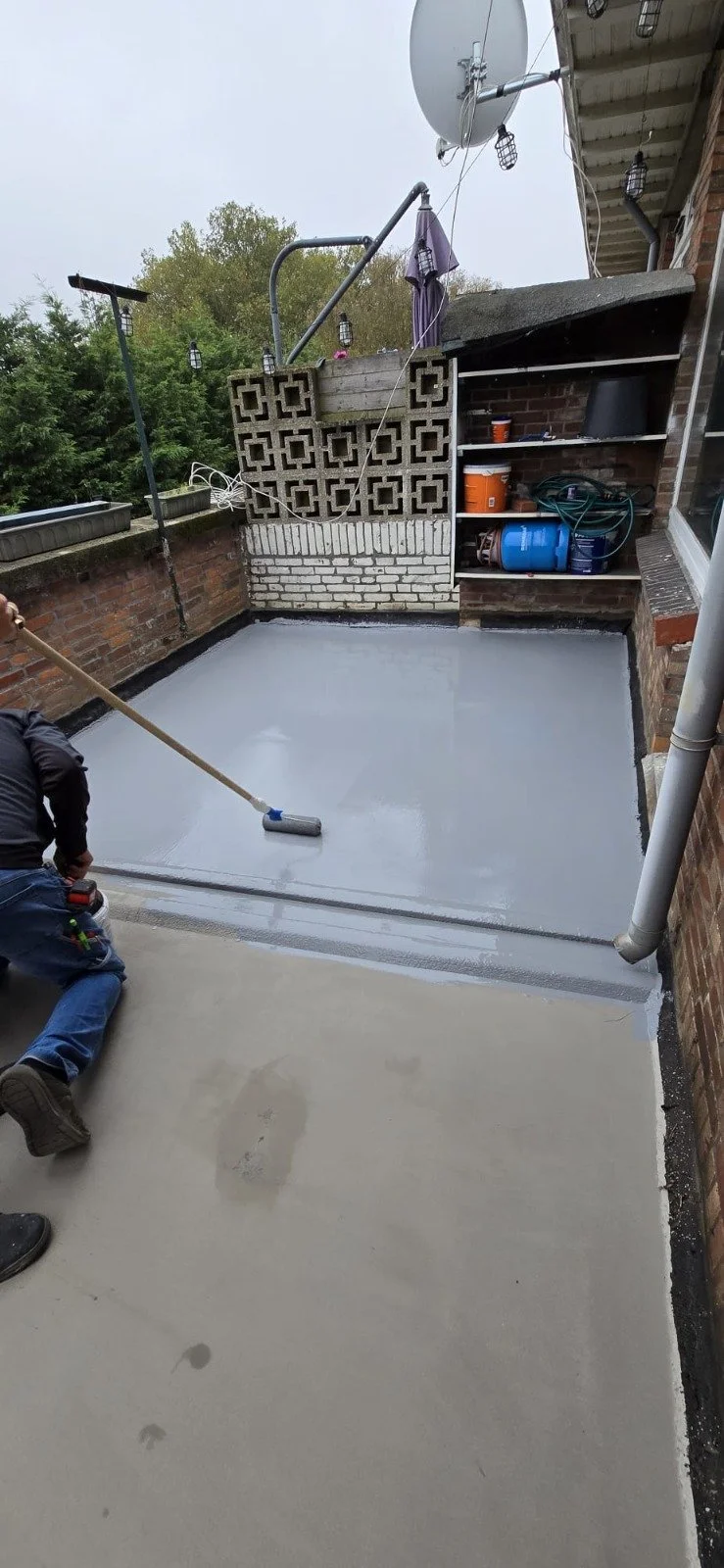 Een persoon is bezig met het uitrollen van een grijze vloercoating op een balkon met bakstenen muren en fietsenrek. Er staan opslagplanken met gereedschap en materialen, en er hangen satellietschotels.