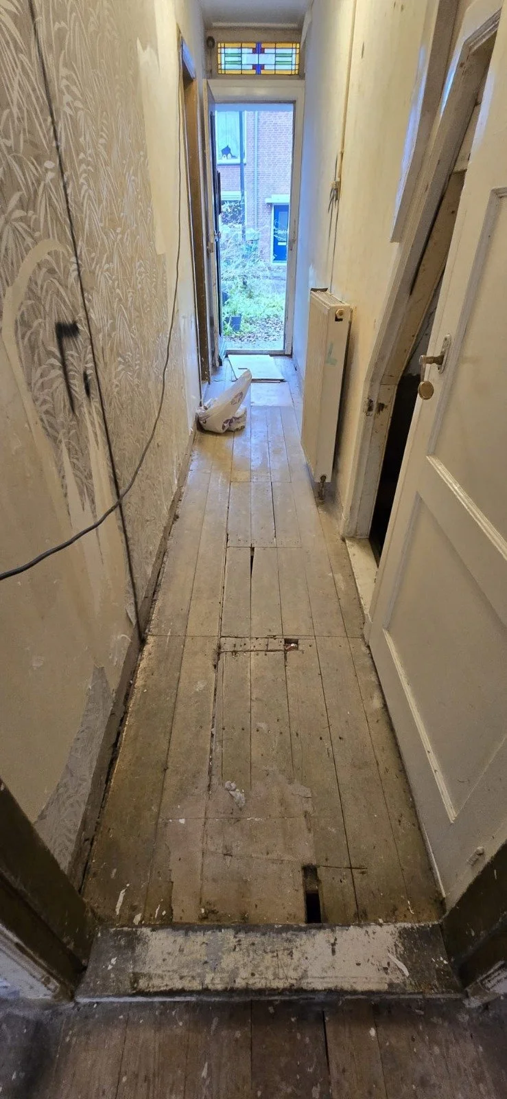 Binnenkijk in een oude, verbouwingswaardige gang met houten vloer die deels is verwijderd, witgekalkte muren, een deur naar buiten, en een glas in lood raam boven de deur.