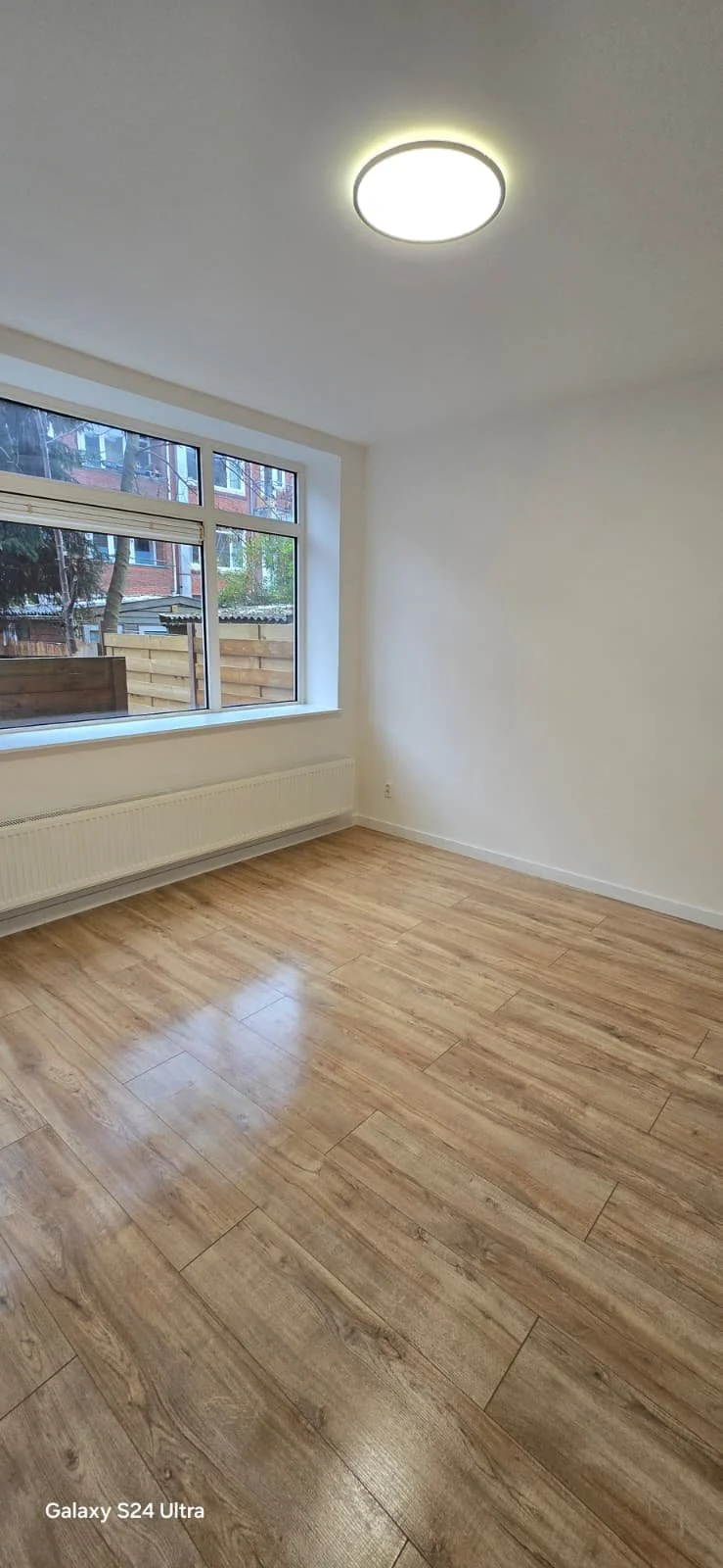 Leeg kamertje met grote ramen, houten vloer en plafondlamp.