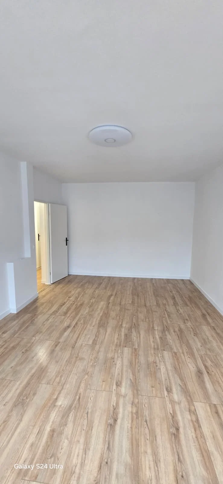 Leeg wit kamertje met houten vloer, white muren, plafondlamp en open deur naar een andere kamer.