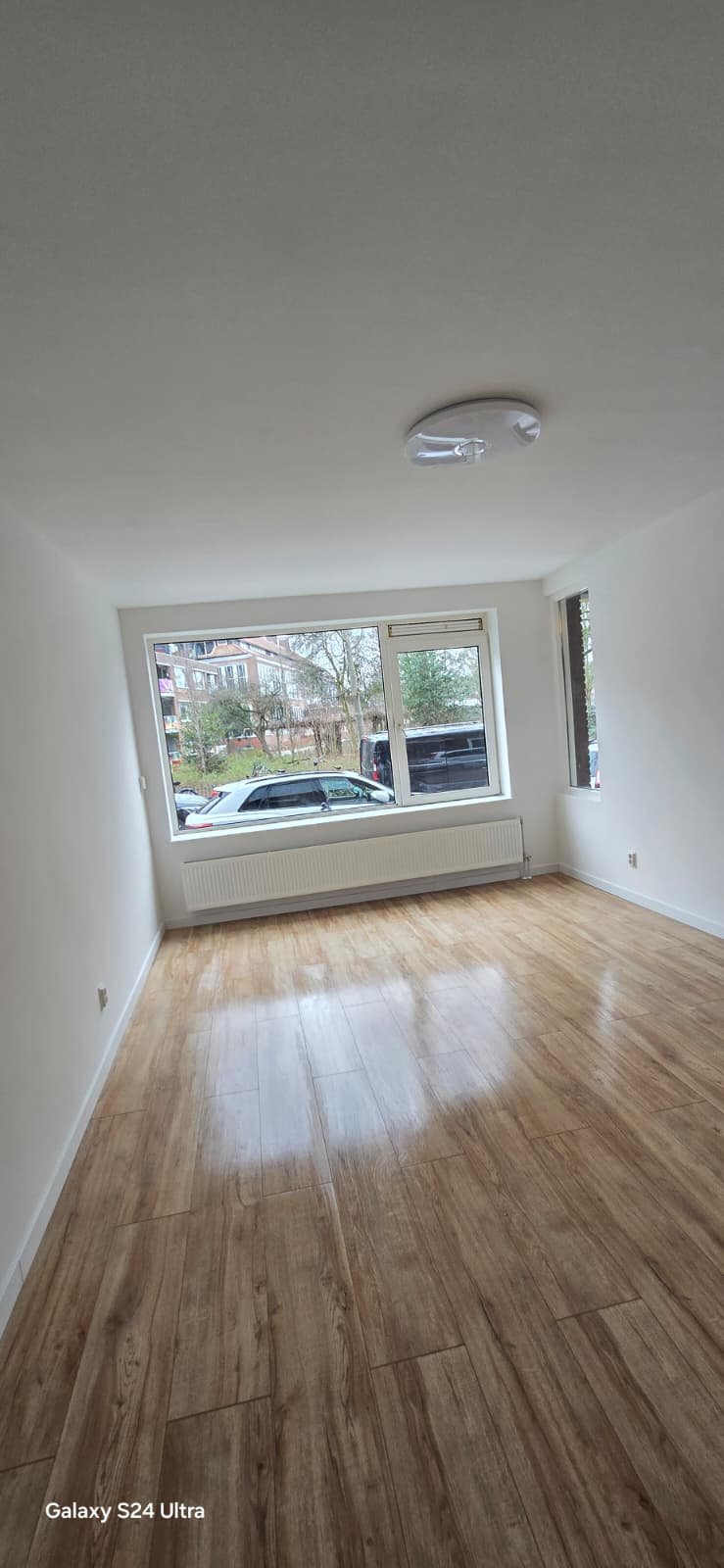 Leeg wit kamertje met grote ramen, houten vloer, radiator onder ramen en plafondlamp.