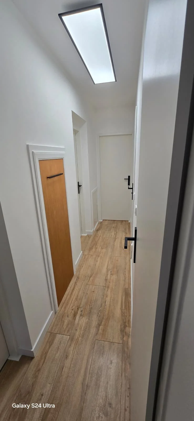 Een gang met witte muren, houten vloeren, drie gesloten witte deuren met zwarte handgrepen en een ingebouwde kast met een houten deur. Een rechthoekige plafondlamp hangt boven.