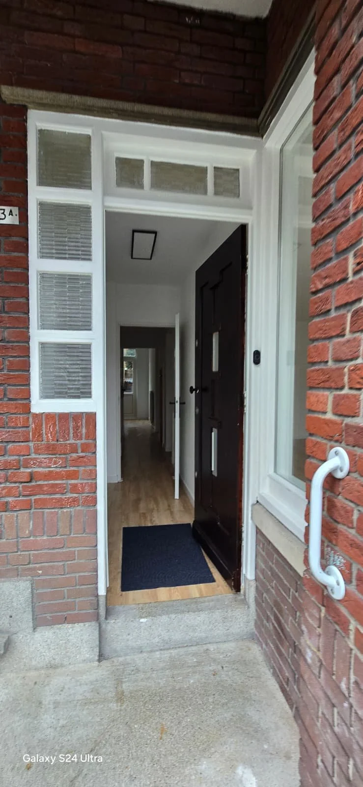De ingang van een huis met een open deur, een mat op de deurmat, zicht op een gang met houten vloer en witte muren, en ramen met jaloezieën. Er is een handslinger aan de muur naast de deur.
