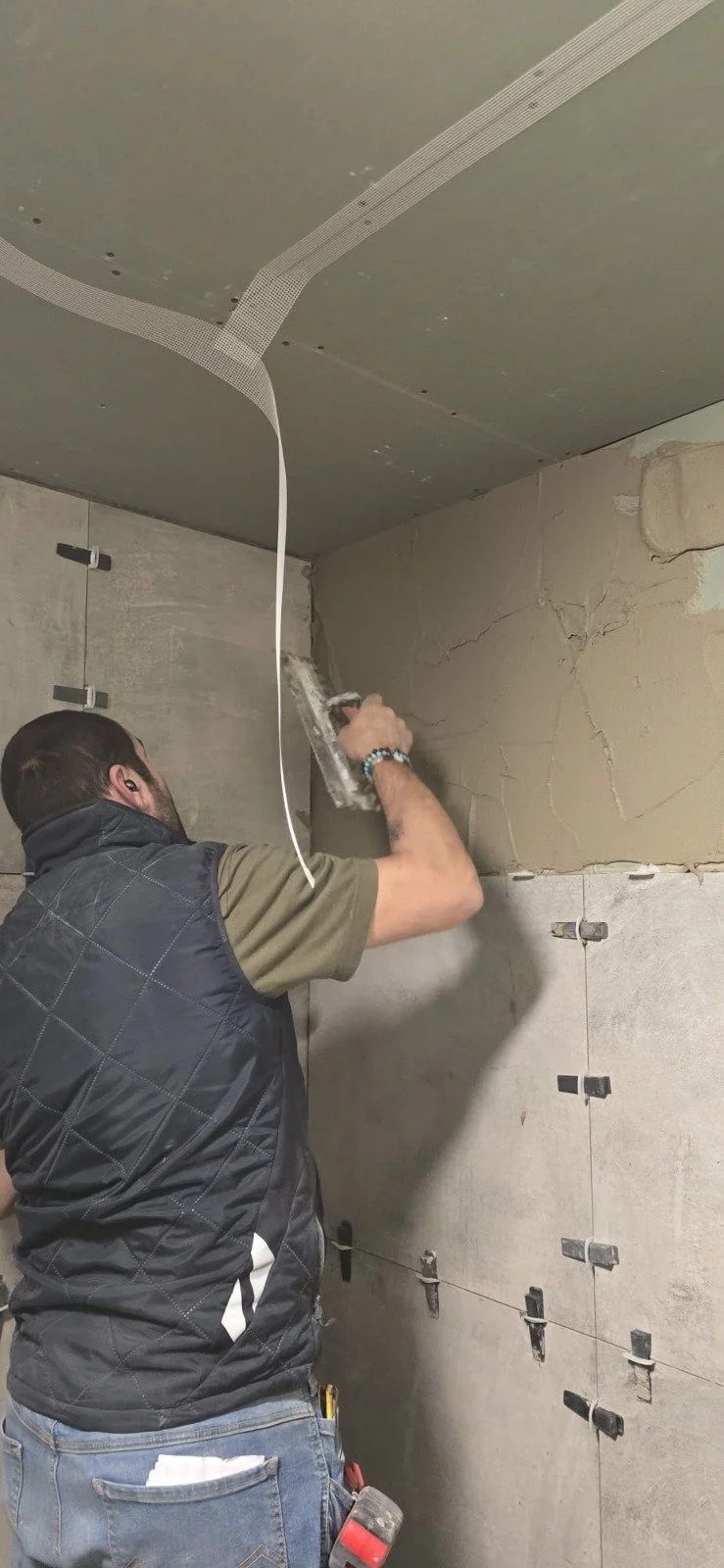 Man die werkzaamheden uitvoert aan de muur, mogelijk het aanbrengen van lijm voor tegels of het behandelen van de muur in een bouw of renovatie project.