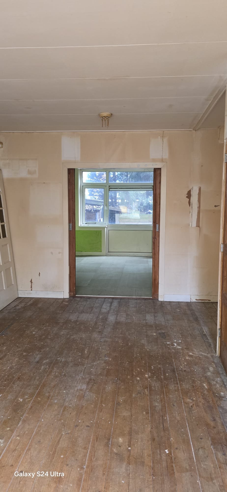 Leeg kamertje met houten vloer en een deur die naar een andere ruimte leidt. De muren en het plafond zijn lichtgekleurd, en er is een verouderde lamp aan het plafond. In de andere ruimte is een grote raam- en deurpartij met uitzicht op buiten.