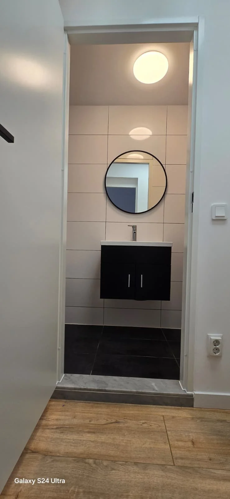 Kleine badkamer met spiegel, wastafel en zwarte wastafelmeubel, betegelde muur, plafondverlichting, schakelaar en stopcontact