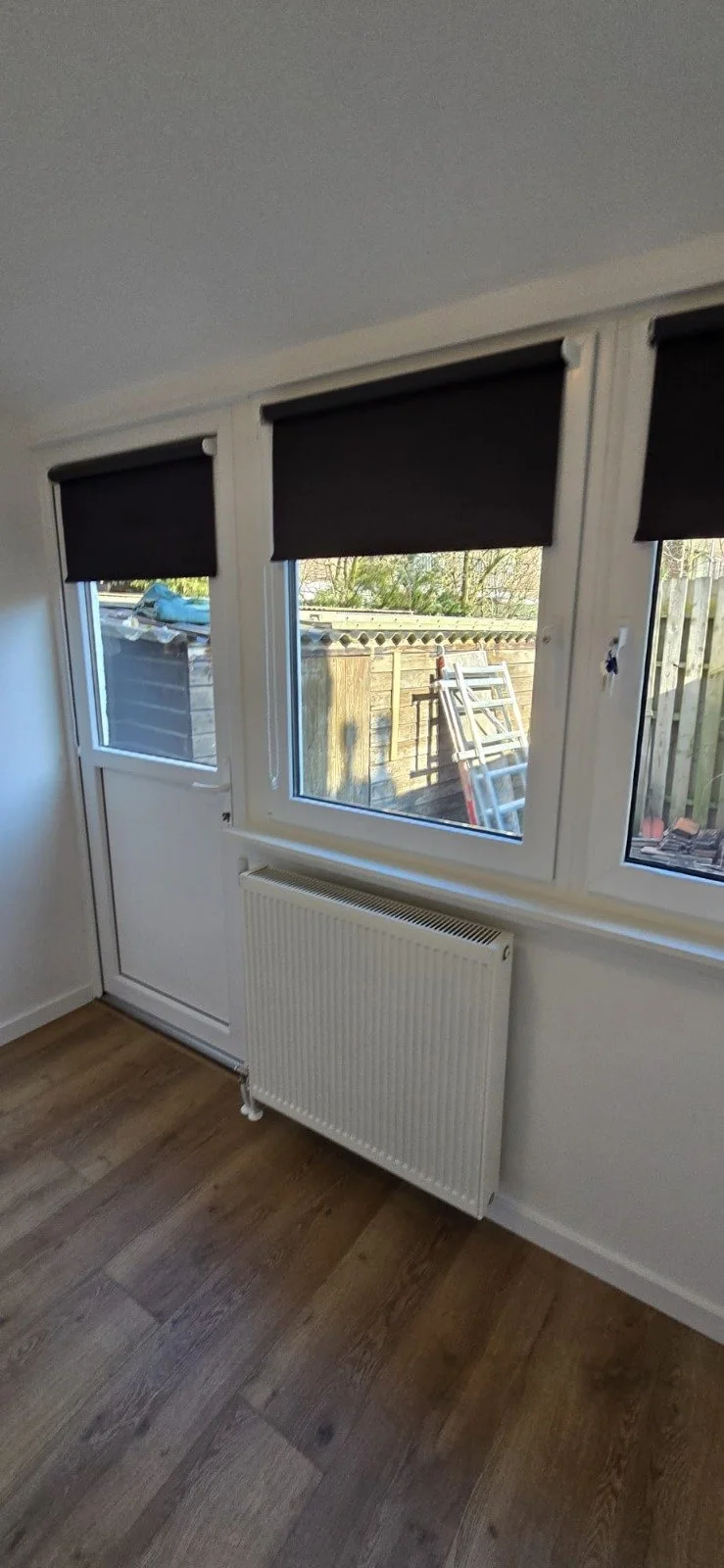 Een binnenruimte met een witte radiator onder een groot raam met drie delen. Het raam bedekt een deur en twee grote ramen. Het raam heeft zwarte rolgordijnen. Buiten is een tuin met een houten schutting en een ladder te zien.