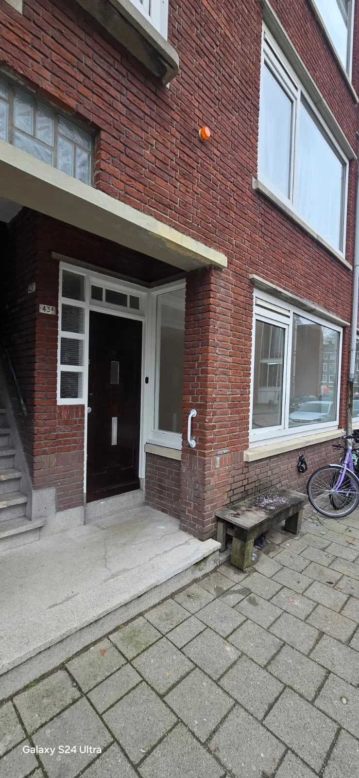 Een rood bakstenen gebouw met meerdere ramen, een witte voordeur en een trap aan de linkerkant. Er ligt een houten bankje voor het gebouw en een fiets aan de rechterkant.