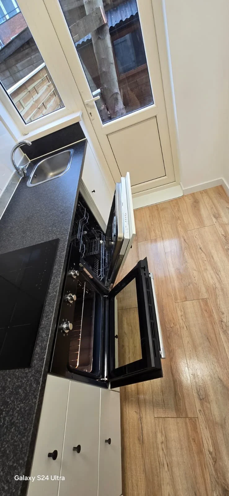 Keuken met zwart werkblad, ingebouwde inductiekookplaat, klein keukensinkje, open oven, witte kasten, deur naar buiten met raam erboven, houten vloer.