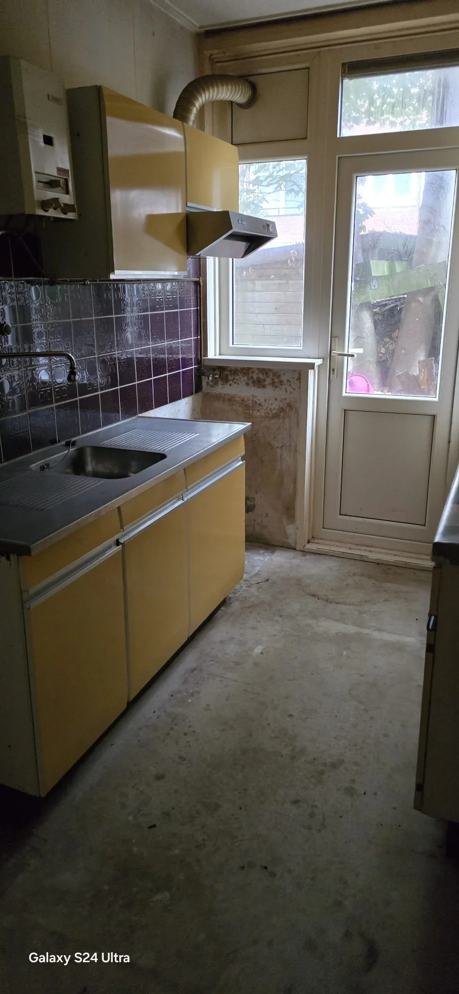 Een oude keuken met gele kasten, een roestige rvs gootsteen, een donkerbruine tegelwand en een witte deur met raam die naar buiten leidt. De vloer is betonnen en gerestaureerd.