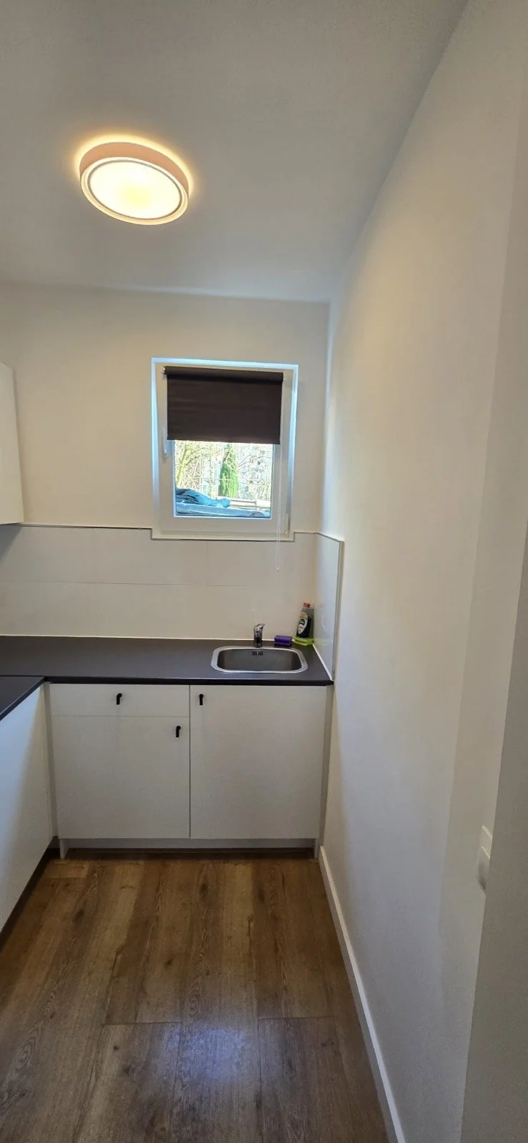 Kleine keuken met wit kast en raam met donker gordijn, kast en vloer van hout, plafondlamp, etensmiddelen op aanrecht.