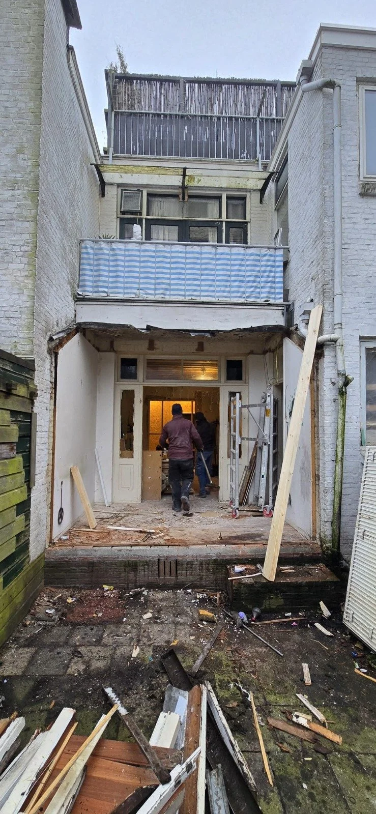 Twee mensen die een verbouwing of renovatie uitvoeren aan de achterzijde van een gebouw, met bouwmaterialen en gereedschap op de vuist.