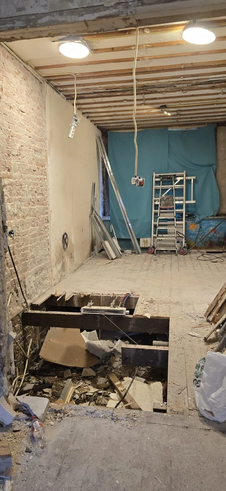 Binnenruimte van een gebouw in aanbouw of renovatie, met een open kelder, ladders, bouwmaterialen en gereedschap.