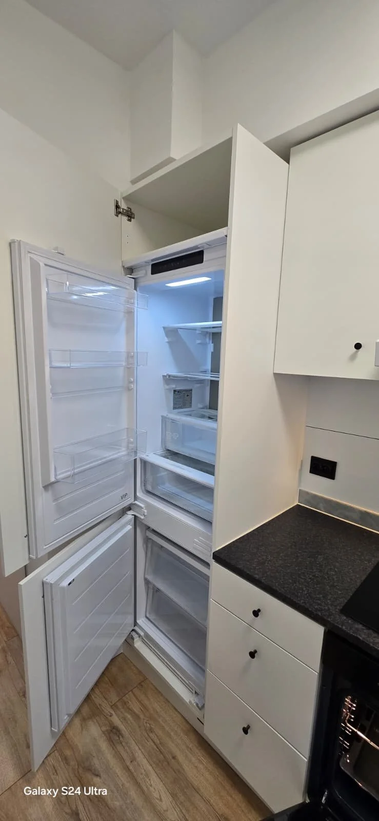 Lege ingebouwde koelkast met open deuren in een witte keuken met zwarte werkblad.