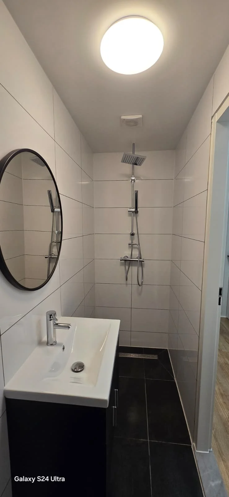 Kleine badkamer met witte tegels, een ronde spiegel, wastafel met zwarte onderkast, douche met regenhanddouche, plafondlamp, en een luchtventilator.
