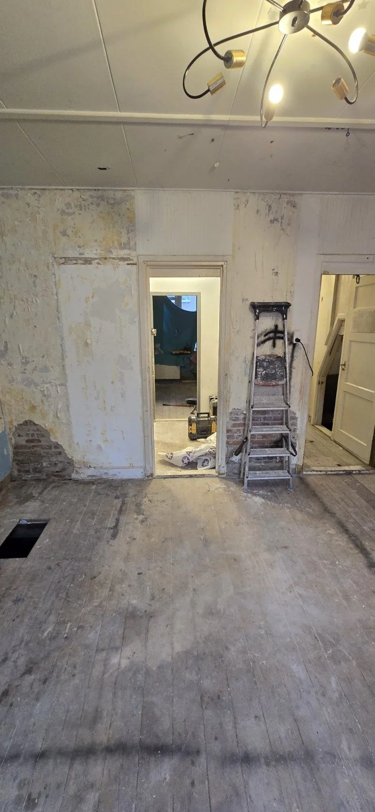 Een kamer in renovatie met een verouderde muur, een ladder en een open deur naar een andere kamer.