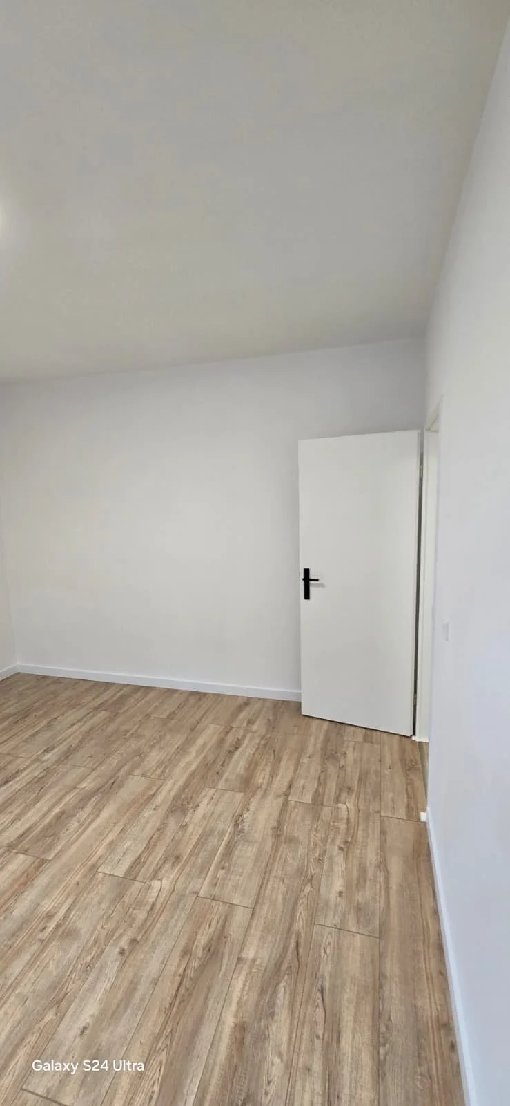 Lege kamer met witte muren, houten vloer en een witte deur met zwarte hendel.