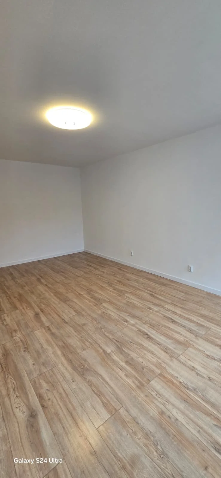 Leeg kamertje met witte muren en houten vloer, verlicht door plafondlamp